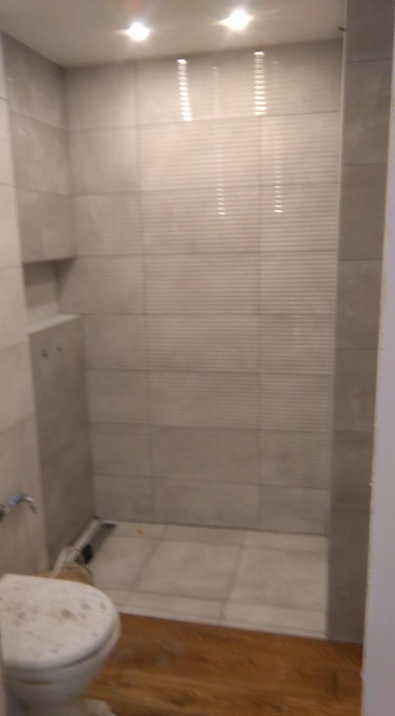 Wykończona łazienka z szarymi płytkami na ścianach i podłodze, widoczny fragment toalety z zabrudzoną deską, oświetlenie sufitowe i zabudowa podtynkowego systemu spłukiwania.