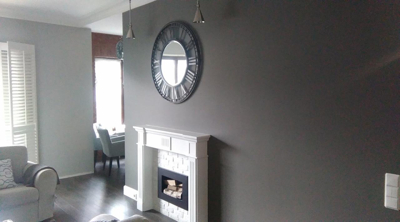 Szary salon z dekoracyjnym kominkiem obłożonym białą cegłą, nad nim okrągłe lustro w stylu zegara, oraz widoczne elementy wystroju wnętrza, takie jak lampy wiszące i meble.