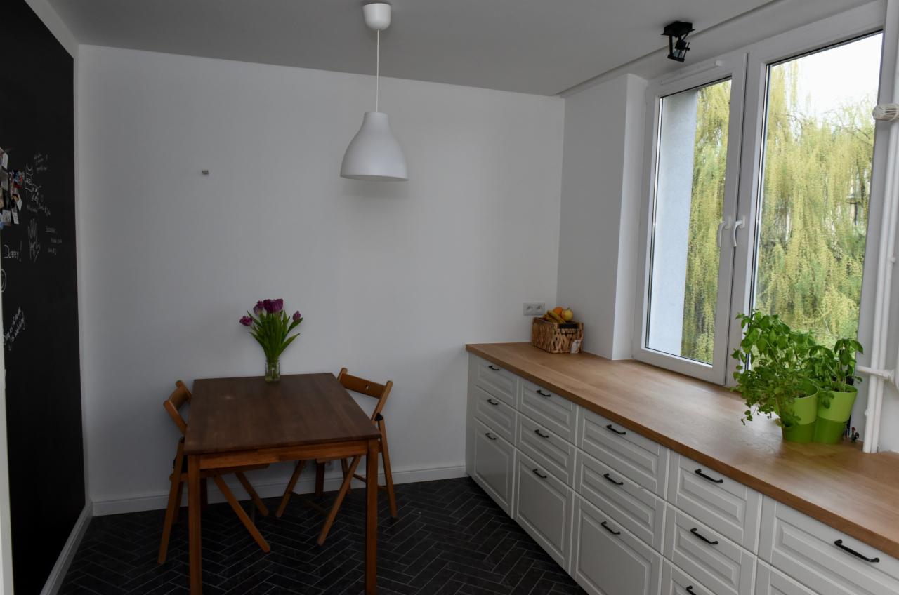 Jasna kuchnia z drewnianym blatem, białymi szafkami z czarnymi uchwytami, drewnianym stołem z fioletowymi tulipanami i czarną ścianą z napisami kredą.