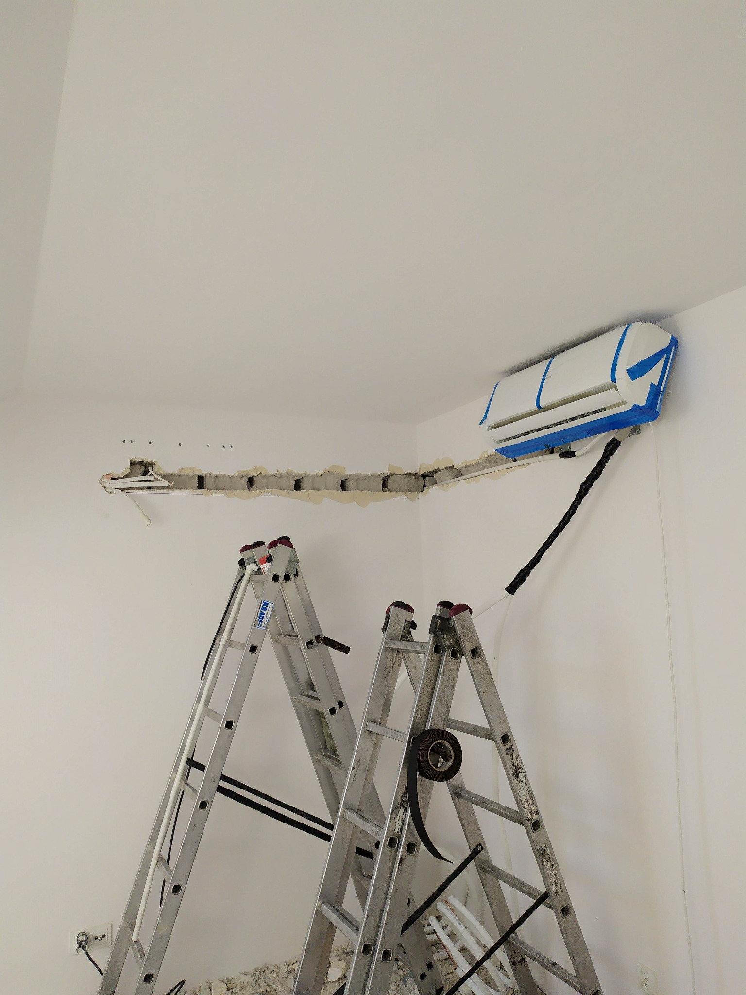 Montaż klimatyzatora w pomieszczeniu, widoczne dwie drabiny aluminiowe, jednostka wewnętrzna klimatyzacji zabezpieczona niebieską taśmą malarską, bruzda w ścianie przygotowana pod instalację.