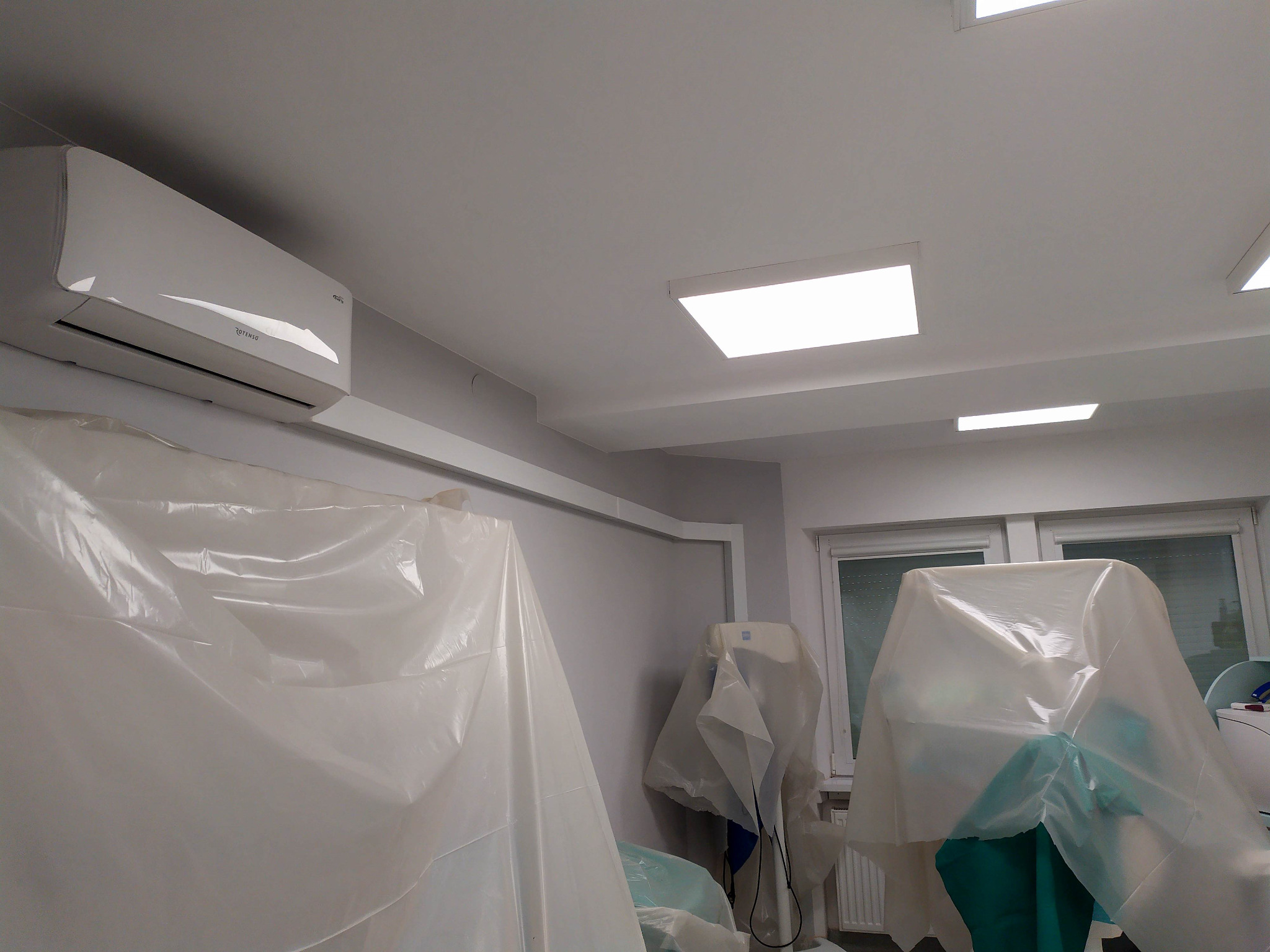 Biały klimatyzator Rotenso zamontowany na ścianie, zabezpieczone folią meble i urządzenia w pomieszczeniu z oknami i panelami LED w suficie.