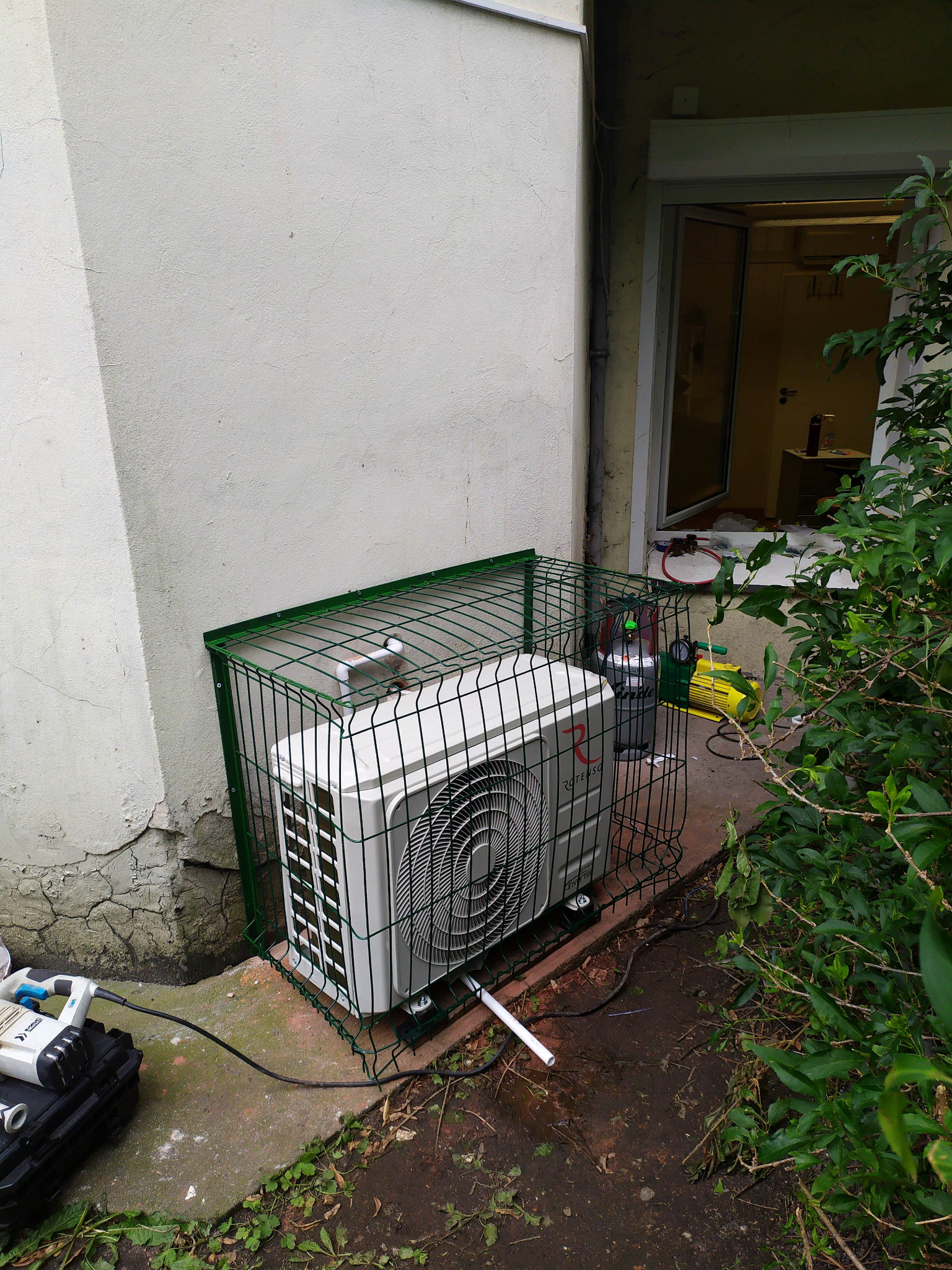 Zewnętrzna jednostka klimatyzatora zabezpieczona zieloną klatką w Szewcach, widoczny sprzęt serwisowy i butla z gazem, ściana budynku z widocznymi spękaniami.