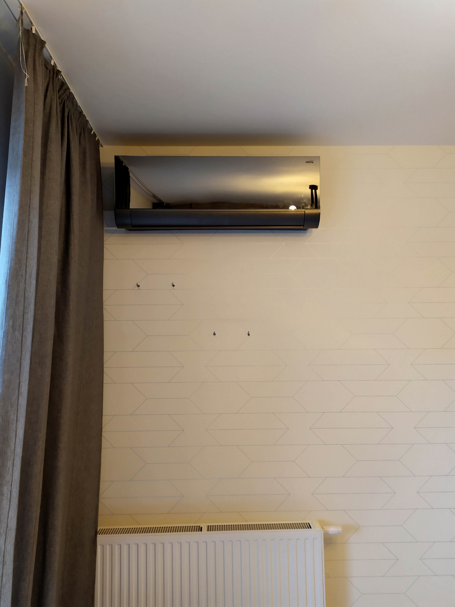 Czarny klimatyzator ścienny zamontowany w pokoju z jasną tapetą w geometryczny wzór, obok zasłony i grzejnika.