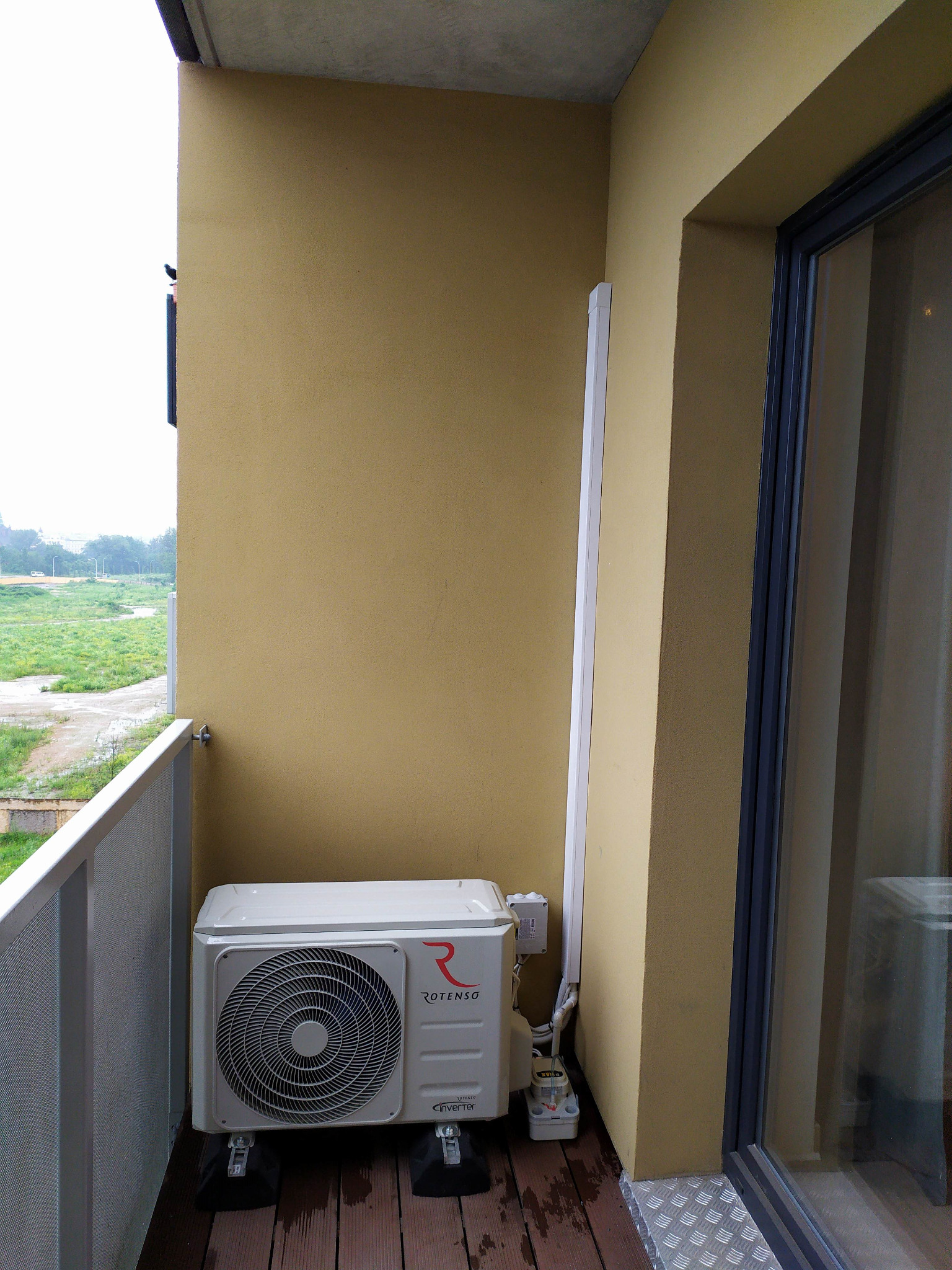 Jednostka zewnętrzna klimatyzacji Rotenso zamontowana na balkonie, widoczne przewody i pompka skroplin, w tle widok na tereny zielone.