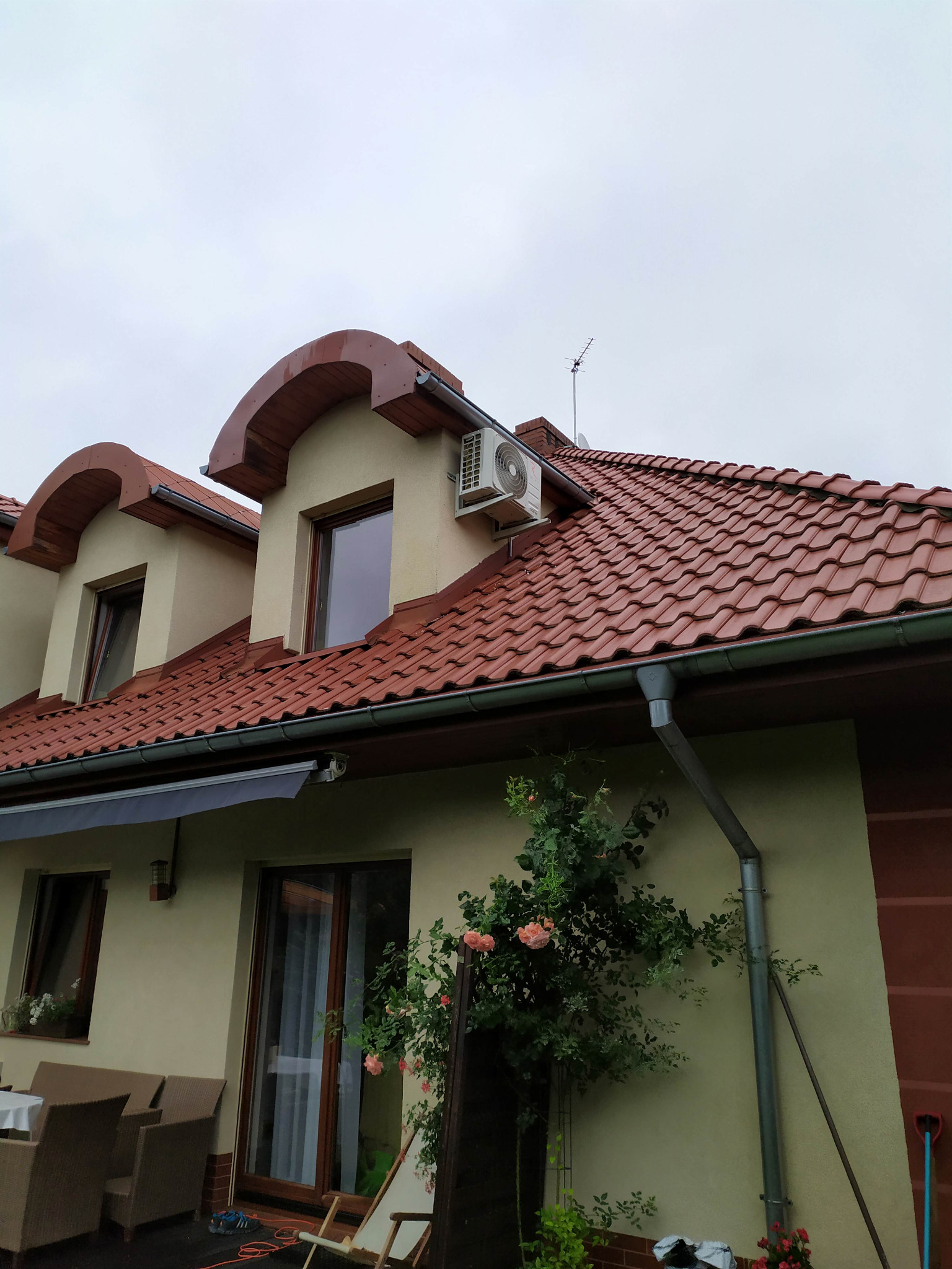 Biała jednostka zewnętrzna klimatyzacji zamontowana na elewacji domu z czerwoną dachówką, widoczna antena telewizyjna na dachu i pnąca się róża przy rynnie.