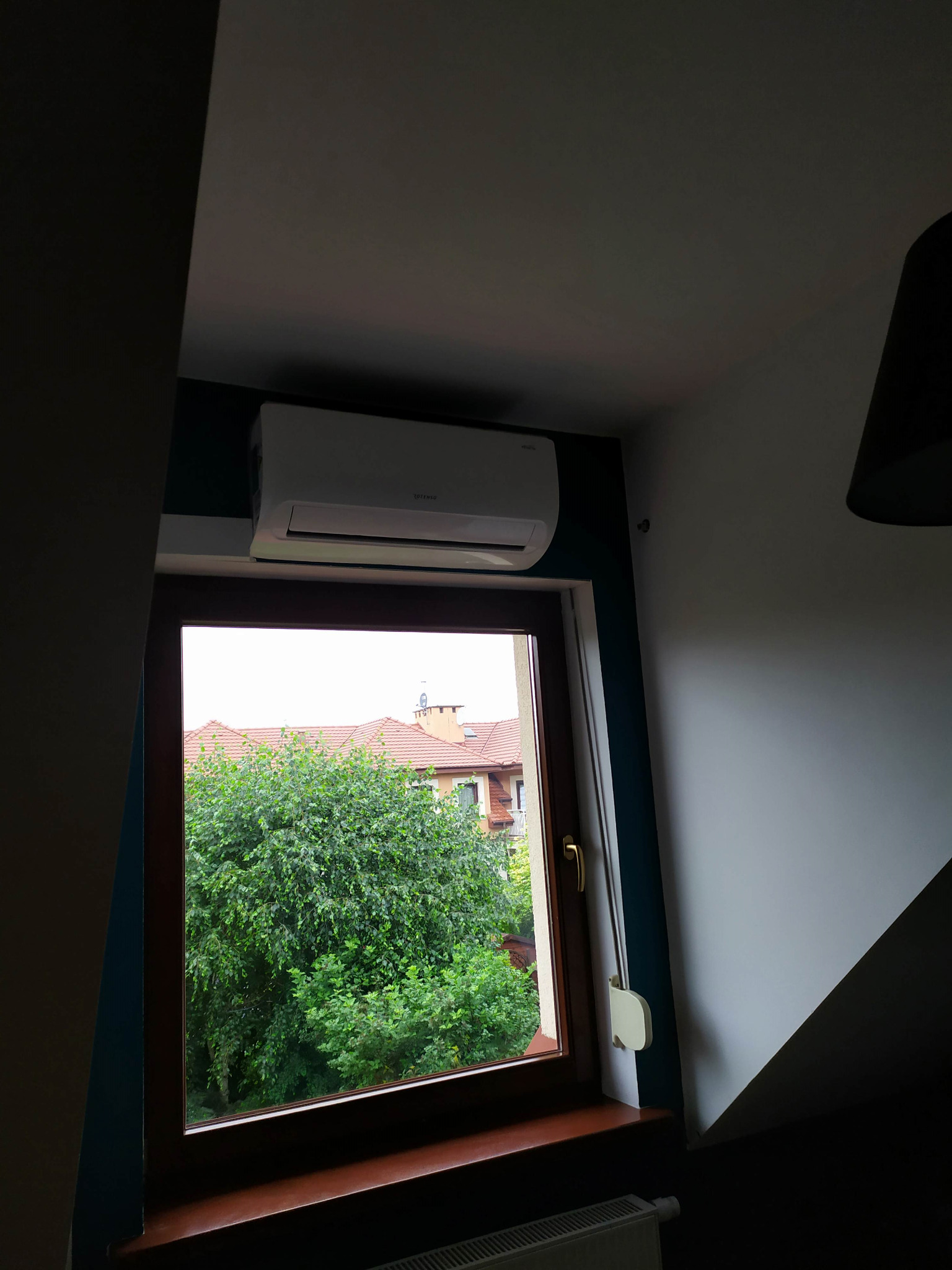 Biały klimatyzator zamontowany nad oknem na poddaszu, widok na zielone drzewa i czerwony dach budynku na zewnątrz.