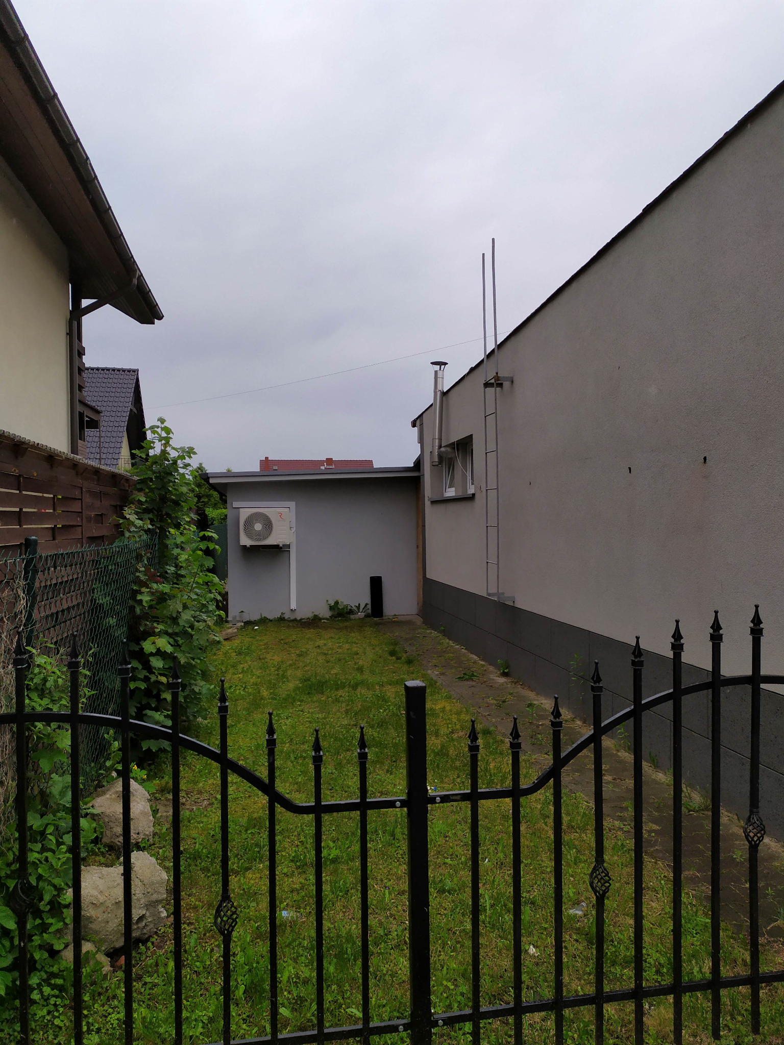 Zewnętrzna jednostka klimatyzacji zamontowana na ścianie szarego budynku gospodarczego, widoczna zza czarnego, metalowego ogrodzenia na tle trawnika i pochmurnego nieba.