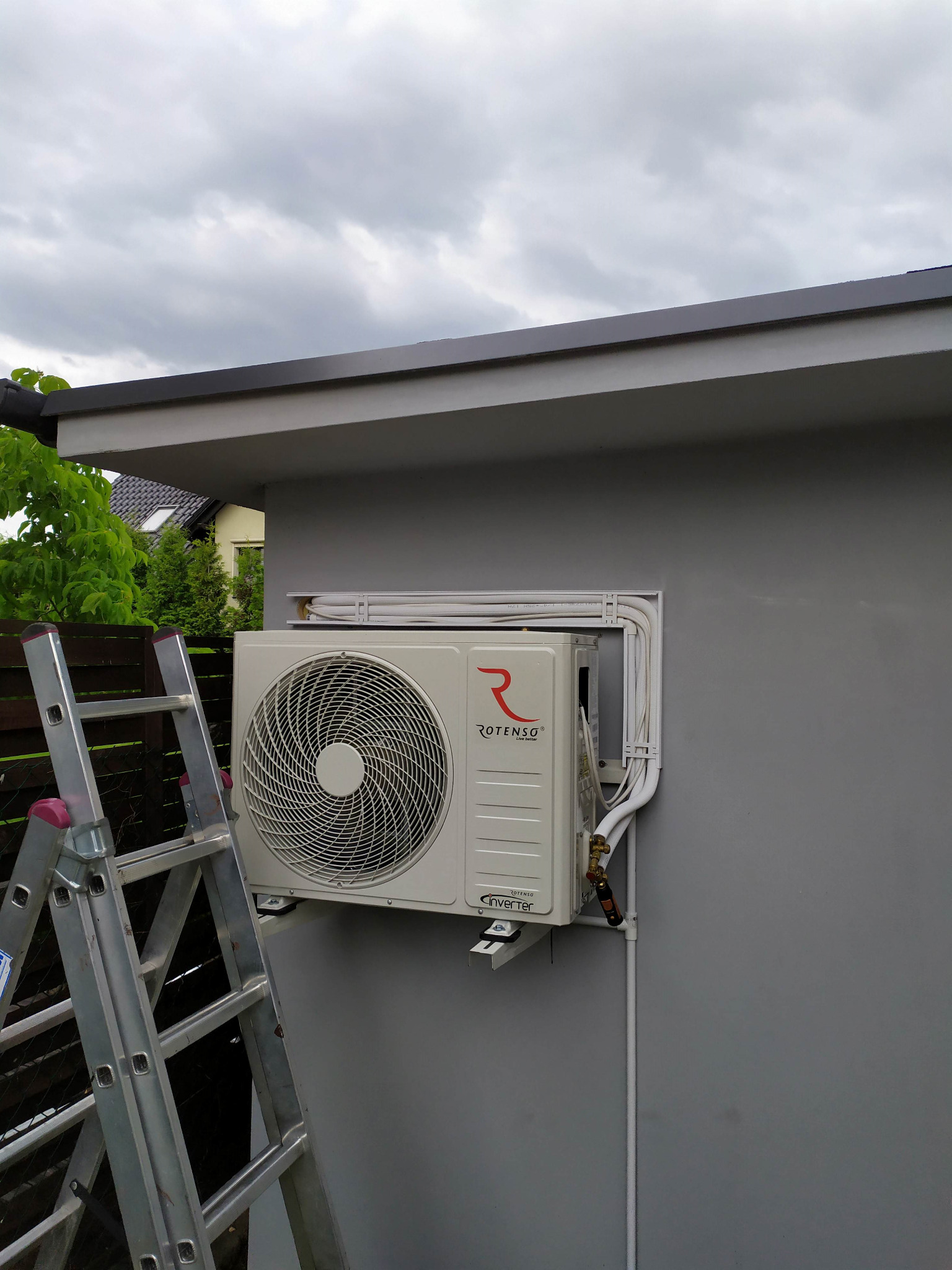 Zewnętrzna jednostka klimatyzacji Rotenso zamontowana na szarej ścianie budynku, z widocznymi rurami w białych maskownicach i drabina przystawna z boku.