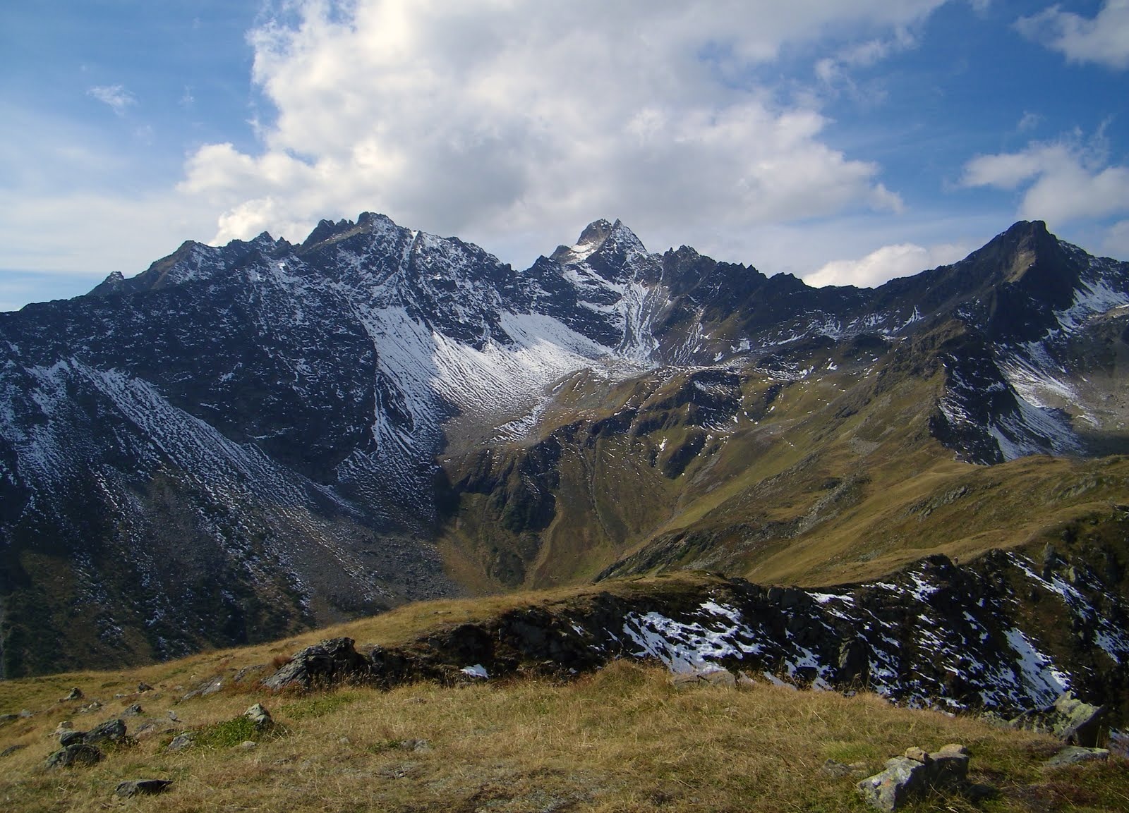 Panoramiczny widok na górzysty krajobraz z ośnieżonymi szczytami pod błękitnym niebem z chmurami, uchwycony z perspektywy trawiastego zbocza.