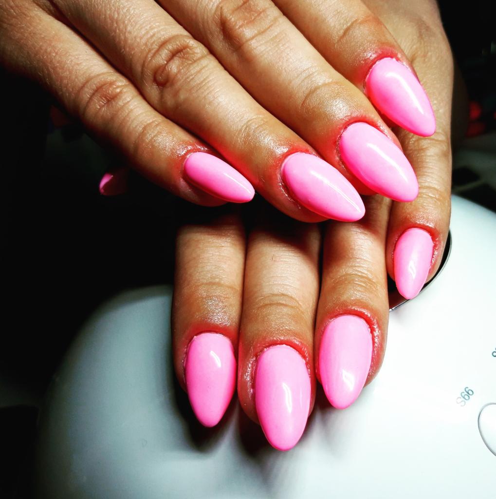 Dłonie z intensywnie różowym, błyszczącym manicure hybrydowym o migdałowym kształcie, widoczne delikatne skórki wokół paznokci, całość na tle lampy UV.
