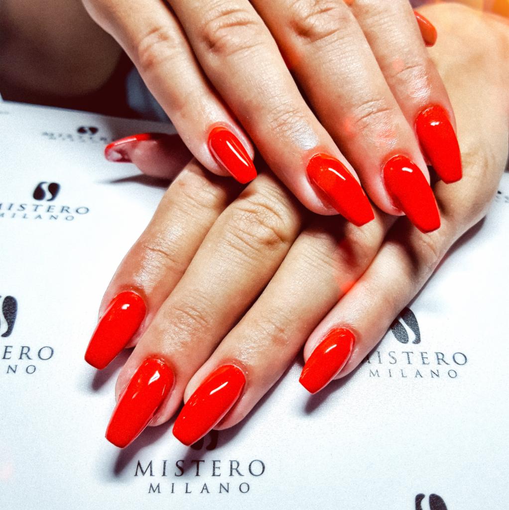 Dłonie z jaskrawoczerwonymi, długimi paznokciami w kształcie migdała, ułożone na białym podkładzie z logo marki kosmetycznej.