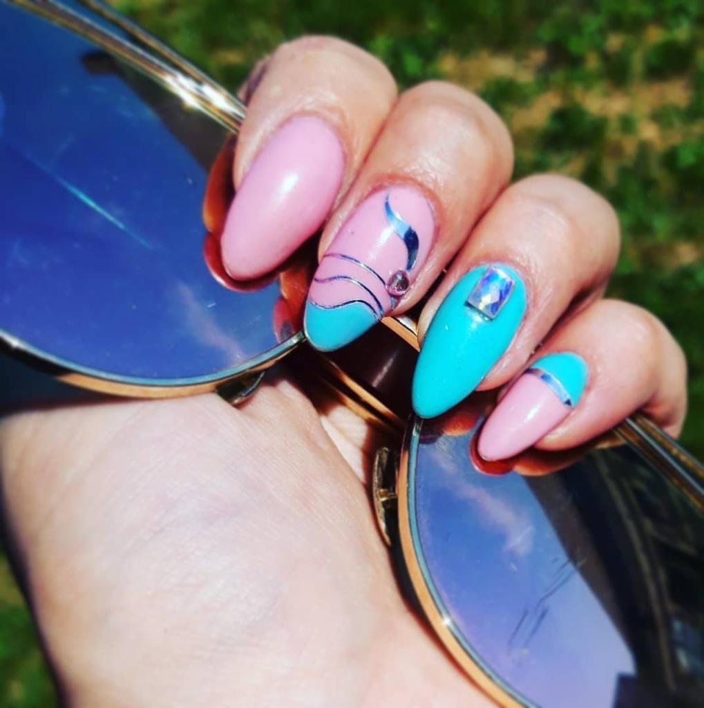 Dłoń z pastelowym manicure hybrydowym w odcieniach różu i turkusu, zdobionym srebrnymi paskami, cyrkoniami i folią, trzymająca okulary przeciwsłoneczne z odbijającym się niebem.