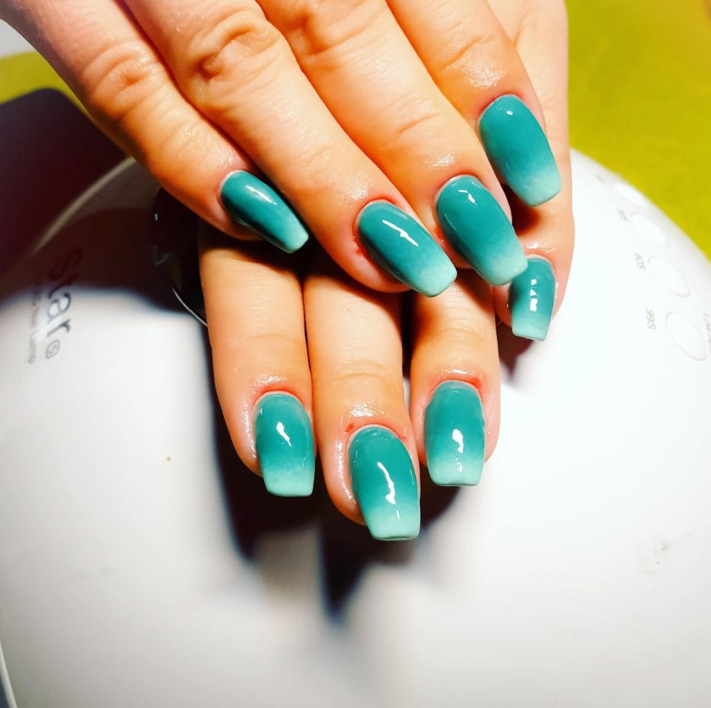 Dłonie z pomalowanymi na turkusowo paznokciami z efektem ombre, jaśniejącym ku końcom, na tle lampy UV do utwardzania lakieru.