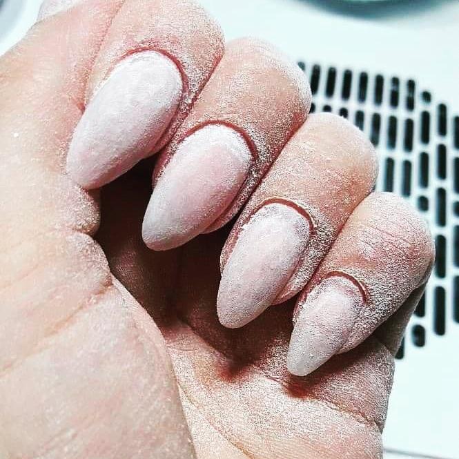 Dłoń z przedłużonymi, pudrowo-różowymi paznokciami w trakcie obróbki, pokryta pyłem po piłowaniu. Widoczny fragment pochłaniacza pyłu.