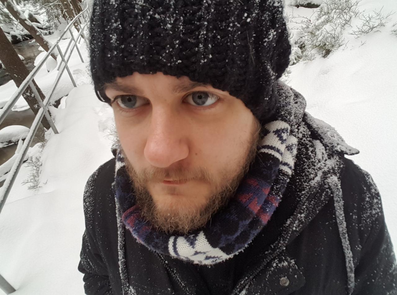 Mężczyzna z brodą w czapce i szaliku w zimowym krajobrazie, pokryty śniegiem