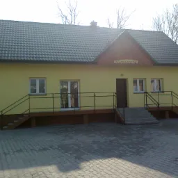 Żółty budynek z ciemnoszarą dachówką ceramiczną, widoczne schody wejściowe i barierki, z napisem 'Połanowiczanka' nad wejściem.