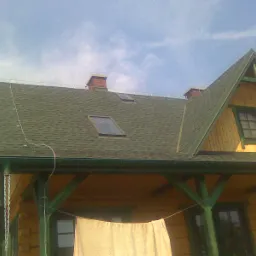 Dom z zielonym dachem pokrytym gontem bitumicznym, widoczne okna dachowe, komin i rynny, na pierwszym planie suszące się pranie.