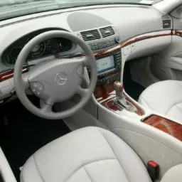 Wnętrze samochodu marki Mercedes z jasną skórzaną tapicerką i drewnianymi elementami wykończeniowymi, widok z przodu na deskę rozdzielczą, kierownicę i fotele.