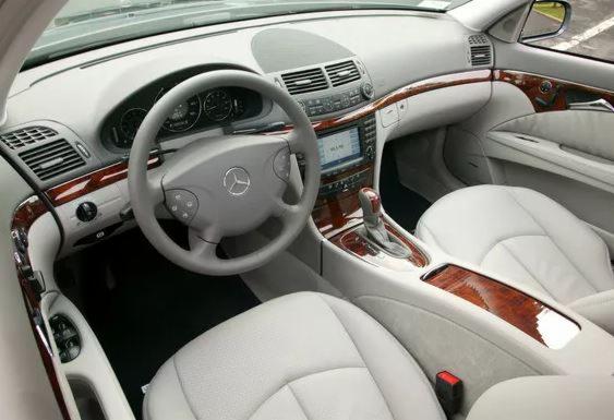 Wnętrze samochodu marki Mercedes z jasną skórzaną tapicerką i drewnianymi elementami wykończeniowymi, widok z przodu na deskę rozdzielczą, kierownicę i fotele.