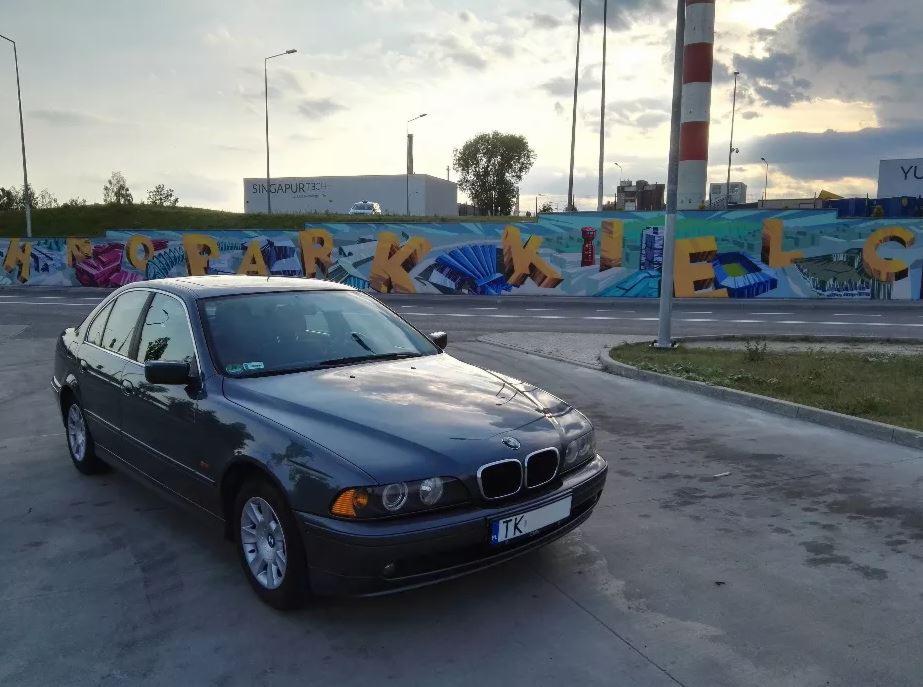 Ciemnoszary sedan BMW E39 zaparkowany na tle kolorowego muralu z napisem 'Kielce', widok z przodu, lekko z boku.