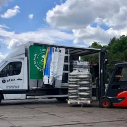 Rozładunek palety z workami z wykorzystaniem wózka widłowego z samochodu dostawczego z logo firmy Atex, w tle błękitne niebo z chmurami i zieleń drzew.