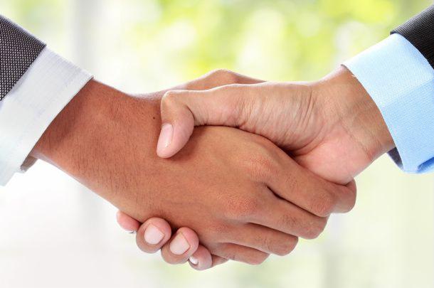 Uścisk dłoni dwóch mężczyzn w garniturach, symbolizujący zawarcie umowy lub partnerstwo biznesowe na tle rozmytego, jasnego tła.