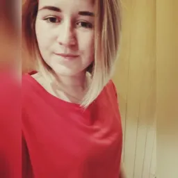 Portret kobiety z blond włosami w czerwonej bluzce na tle drewnianej ściany. Uśmiechnięta twarz skierowana w stronę obiektywu, naturalne światło.