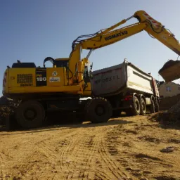 Żółta koparka Komatsu PW180 ładuje ziemię do wywrotki na placu budowy, widoczne ślady opon na piaszczystym podłożu i sterta ziemi w tle.