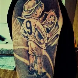 Tatuaż na ramieniu przedstawiający chłopca w stroju piłkarskim z numerem 9, trzymającego piłkę, na tle gór i liścia dębu.
