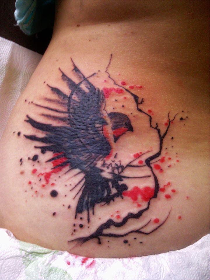 Tatuaż przedstawiający lecącego ptaka z elementami drzewa i czerwonymi plamami, wykonany na skórze w okolicy biodra.