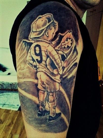 Tatuaż na ramieniu przedstawiający chłopca w stroju piłkarskim z numerem 9, trzymającego piłkę, na tle gór i liścia dębu.