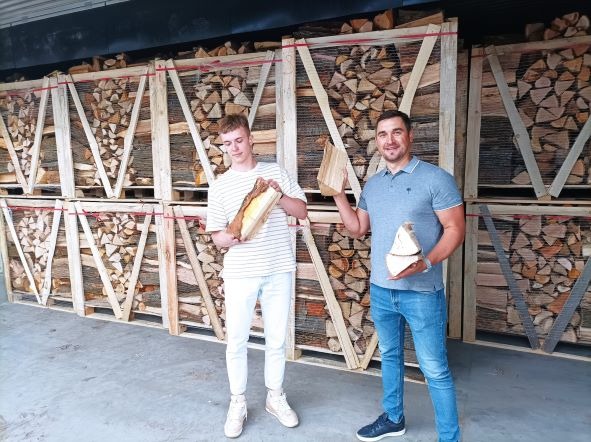 Dwaj mężczyźni prezentują kawałki drewna opałowego na tle palet wypełnionych pociętym drewnem w magazynie.