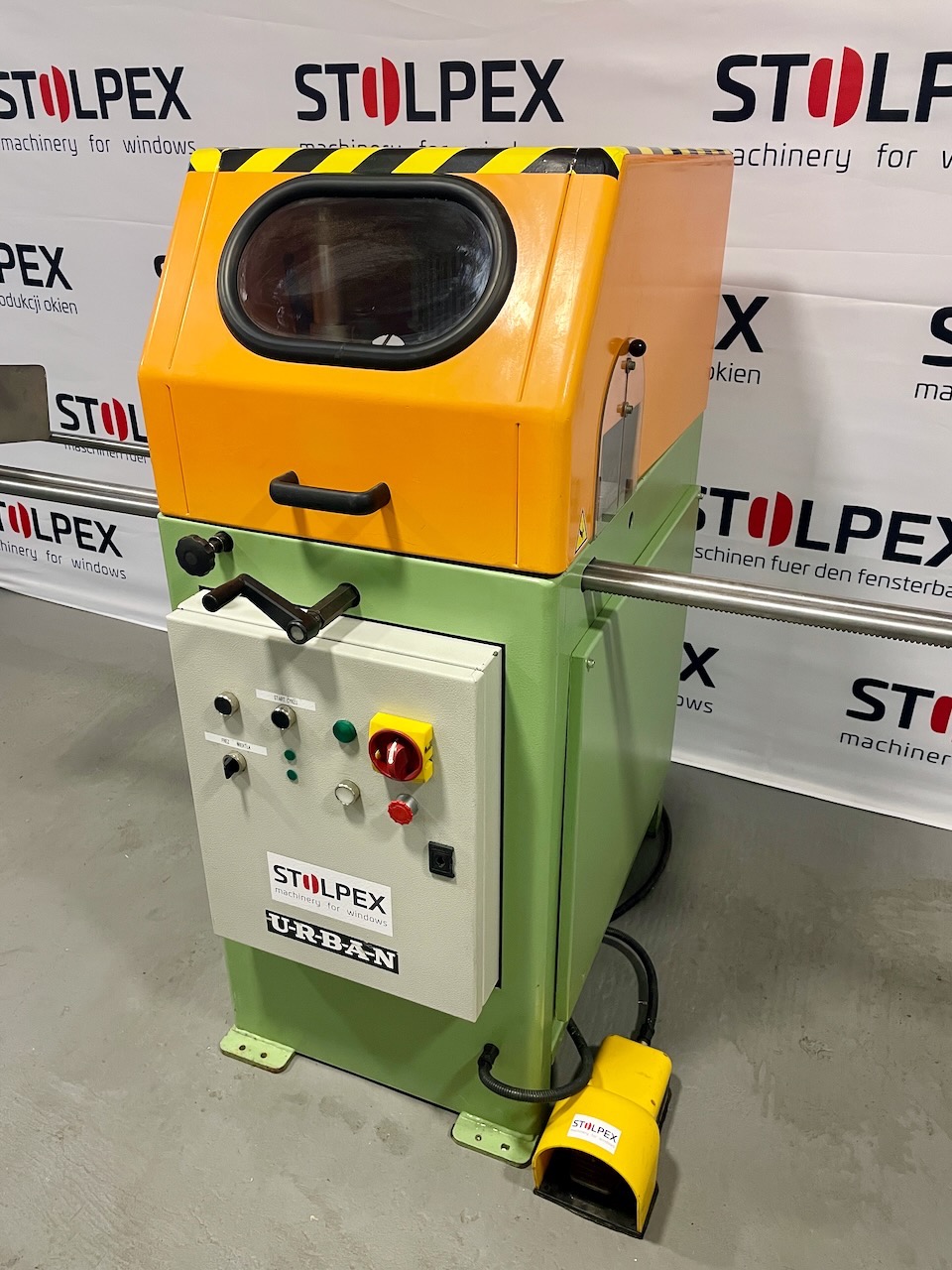Stacjonarna maszyna do obróbki okien z żółto-zieloną obudową, panelem sterowania z przyciskami i żółtym pedałem nożnym, marki Stolpex Urban, na tle z logo firmy.