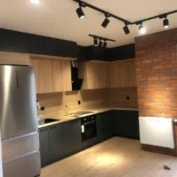 Nowoczesna kuchnia z drewnianymi szafkami g&oacute;rnymi i grafitowymi dolnymi, ceglana ściana z białym grzejnikiem, oświetlenie punktowe na suficie, widok na balkon.