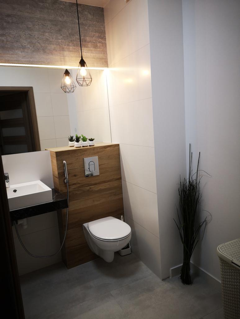 Nowoczesna łazienka z wiszącą toaletą, umywalką na blacie z czarnego granitu i drewnianą zabudową, oświetlona lampami wiszącymi w metalowych kloszach.