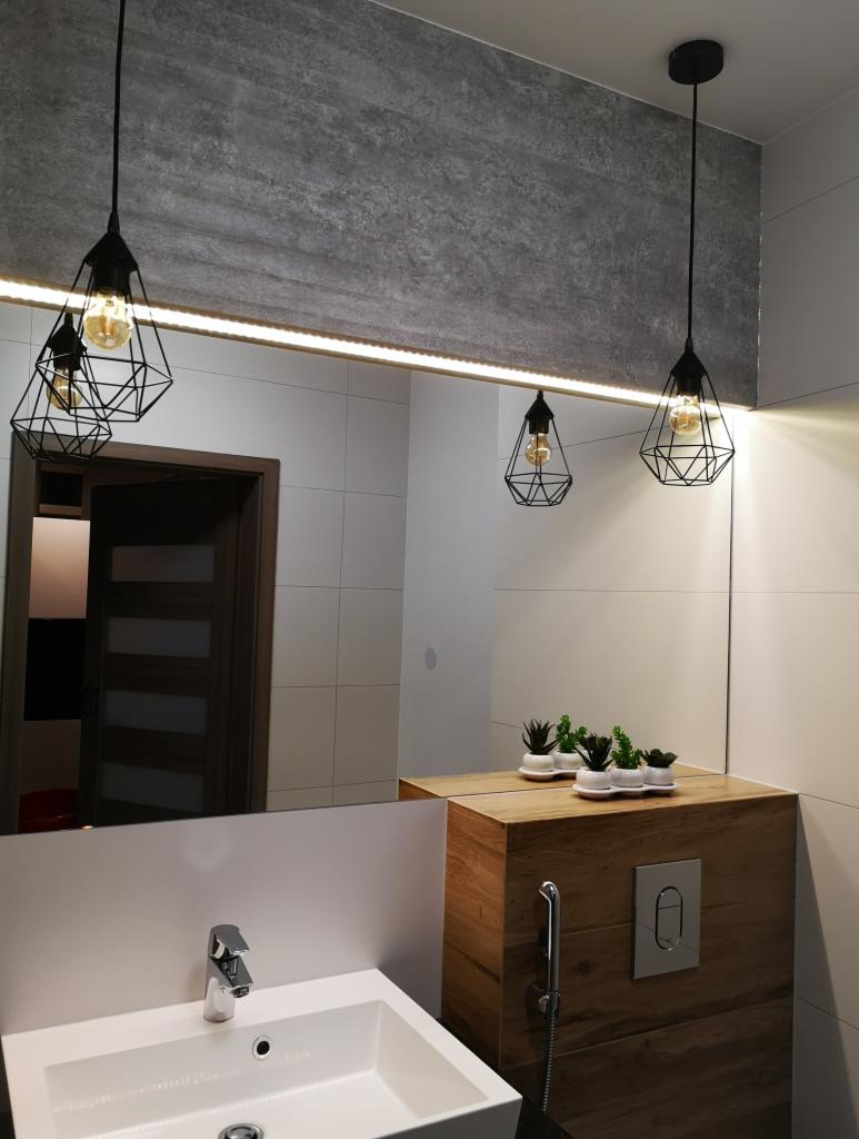 Nowoczesna łazienka z białą umywalką, drewnianą szafką pod umywalką, i trzema wiszącymi lampami w geometrycznych oprawach, odbijającymi się w dużym lustrze z podświetleniem LED.