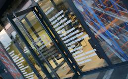 Szklana fasada budynku z widocznymi napisami i odbiciami konstrukcji stalowej wewnątrz, widziana pod kątem.