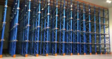 Konstrukcja stalowa, niebieskie słupy i pomarańczowe podstawy tworzące szkielet regału magazynowego w hali.