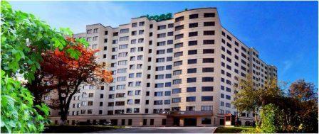 Wielopiętrowy budynek hotelowy z zielonym napisem na dachu, otoczony zielenią i drzewem z czerwonymi liśćmi, widok z zewnątrz w słoneczny dzień.