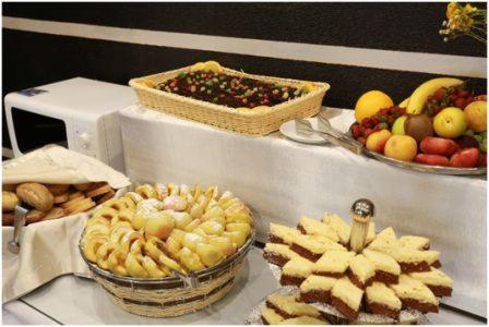 Stół bufetowy z ciastami, pieczywem i owocami, widoczny tort czekoladowy w koszyku, ciasto z owocami w koszyku, talerz z owocami, ciasto w kształcie gwiazdy, pieczywo w koszyku, biały obrus...