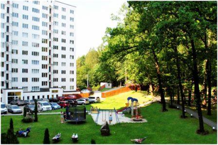 Wysoki apartamentowiec z parkingiem i placem zabaw, otoczony zielenią drzew i trawnikiem.