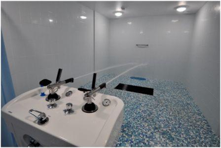 Hydromasaż: biała wanna z dyszami i armaturą, woda pod ciśnieniem, mozaika w odcieniach błękitu na podłodze i ścianach, jasne oświetlenie sufitowe.