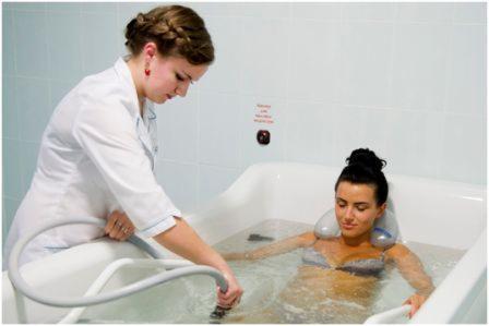 Kobieta w wannie z hydromasażem, obsługiwana przez osobę w białym uniformie, trzymającą rurę do masażu wodnego.