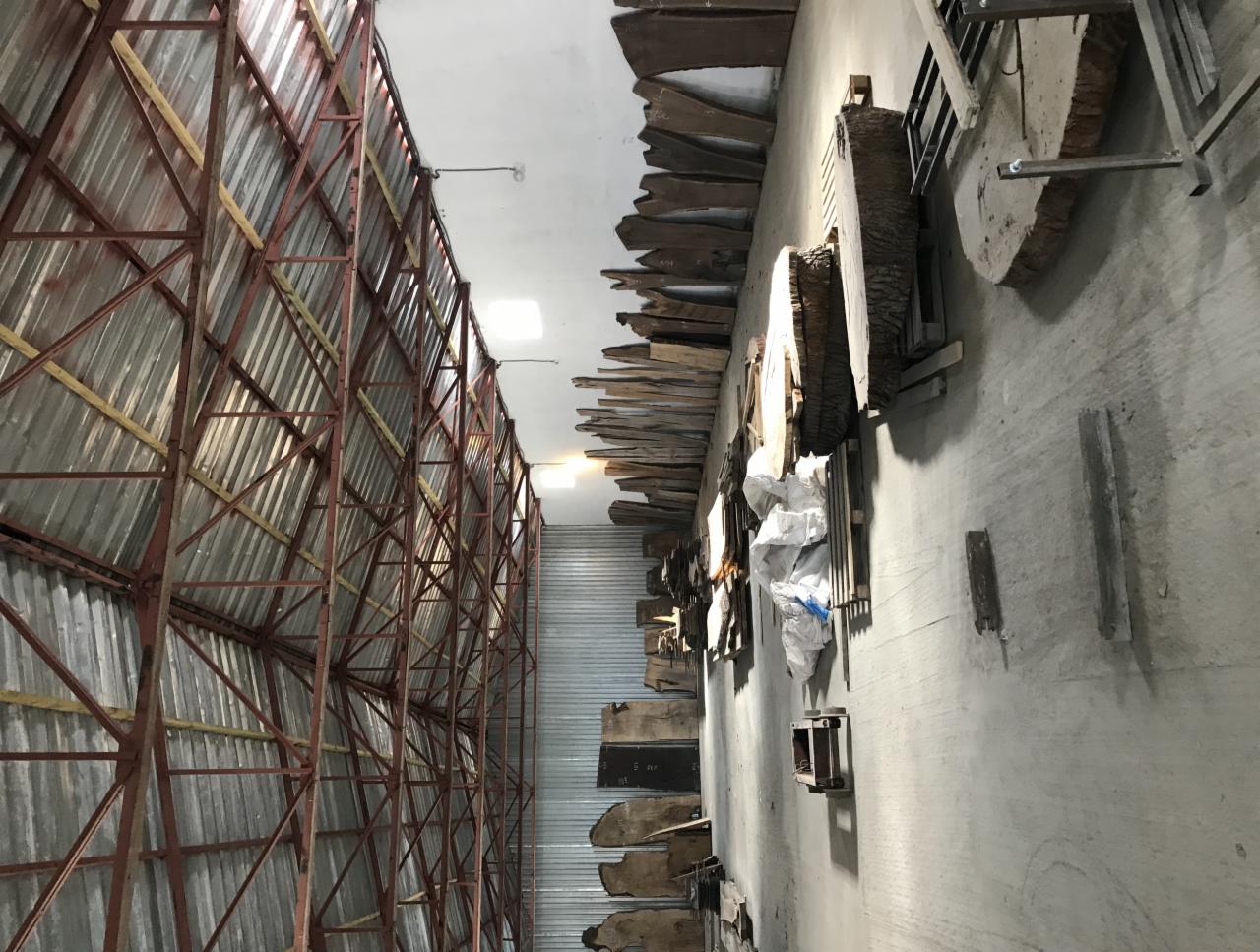 Magazyn z surowym drewnem w plastrach, przygotowanym do obróbki na blaty, deski i inne elementy, widoczne metalowe konstrukcje regałów i oświetlenie.