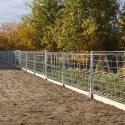 Długi, metalowy płot panelowy z podmurówką, widok z perspektywy ziemi, w tle drzewa z jesiennymi liśćmi.