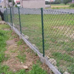 Fragment nowo postawionego ogrodzenia z siatki z zielonymi słupkami i betonową podmurówką, widok na zarośnięty trawnikiem teren działki i sąsiednie budynki.