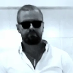 Mężczyzna z brodą i okularami przeciwsłonecznymi w białej koszuli, portret w czerni i bieli.