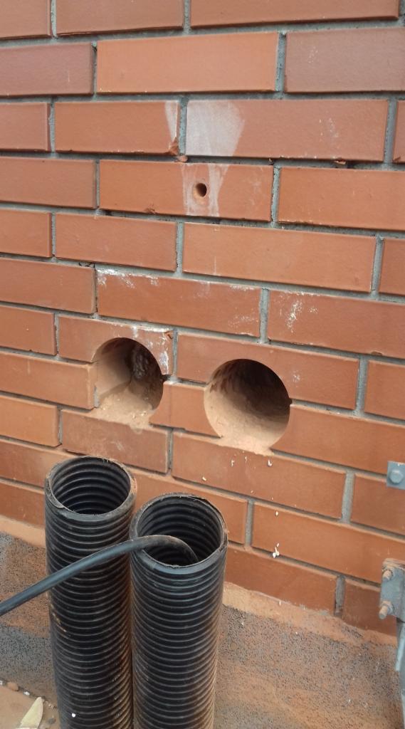 Ceglana ściana z dwoma okrągłymi otworami, poniżej widoczne dwie czarne rury karbowane, ślady po wierceniu i drobne uszkodzenia cegieł w okolicy otworów.