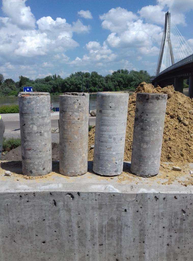 Cztery rdzenie betonowe wywiercone w fundamencie na tle mostu wantowego i błękitnego nieba z chmurami.