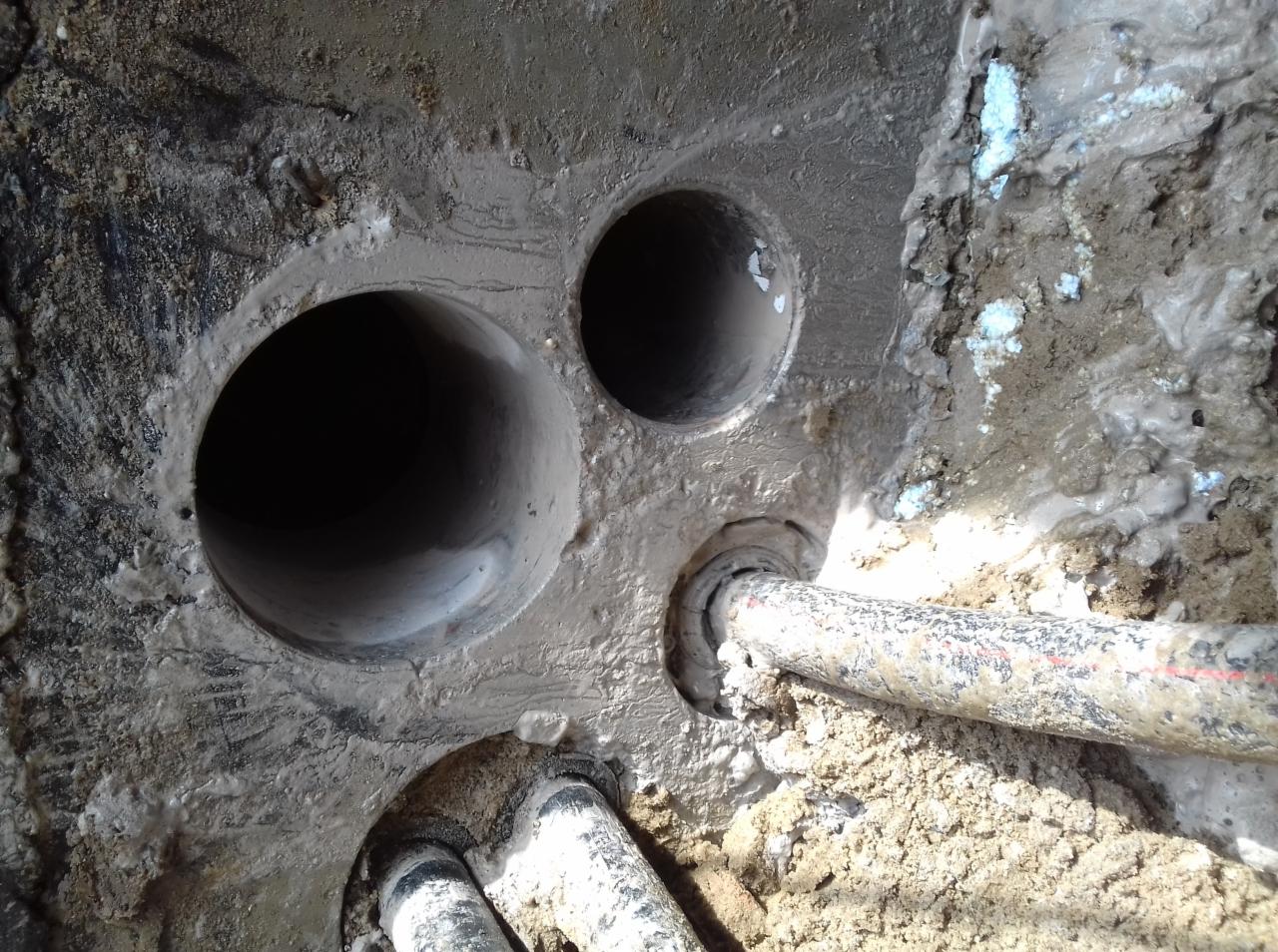 Betonowa ściana z trzema okrągłymi otworami o różnych średnicach, przez jeden z nich przechodzi rura z czerwoną linią, otoczenie zabrudzone zaprawą.